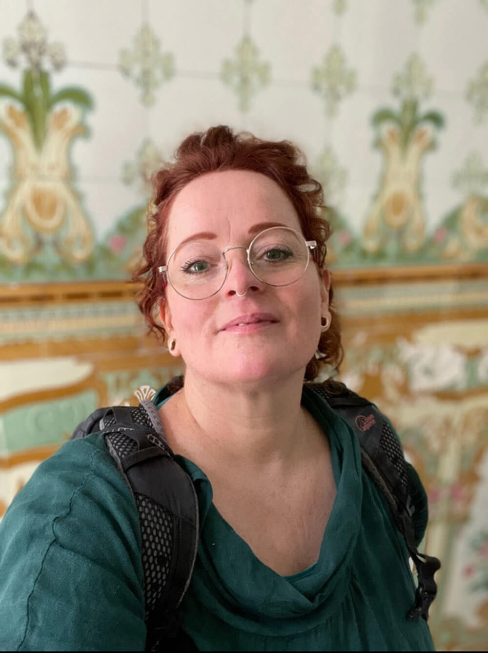 ginny-warner-doula-persoonlijk-verhaal-zorgnomade Ginny Warner, doula bij rouw en sterven, kijkt met zachte blik in de camera voor een sierlijke wand; portret op de pagina Ginnys Verhaal van Zorgnomade.